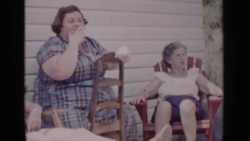 In 1951 New York home-movie footage, the woman in the plaid dress brings a hand toward her mouth, with another woman seated nearby on the porch.