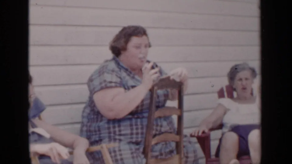 In 1951 New York home-movie footage, a woman in a blue plaid dress appears to smoke beside another seated woman against a clapboard exterior wall.