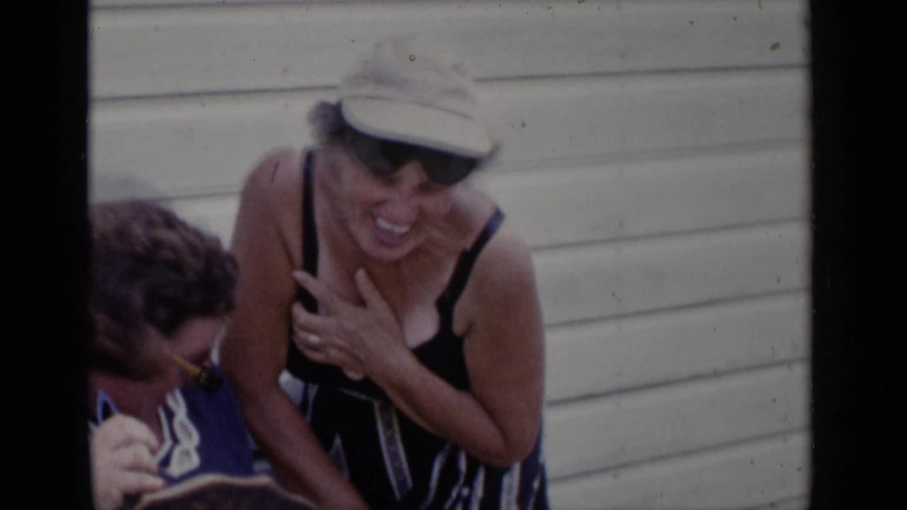 Two older women laugh beside light-colored house siding, one in sunglasses and one in a sun hat.