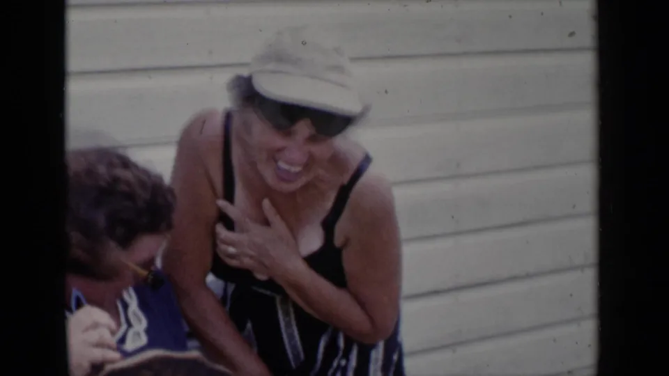 1951 New York 8mm frame of two older women laughing outdoors beside light-colored siding, with one wearing yellow sunglasses and the other a pale hat.