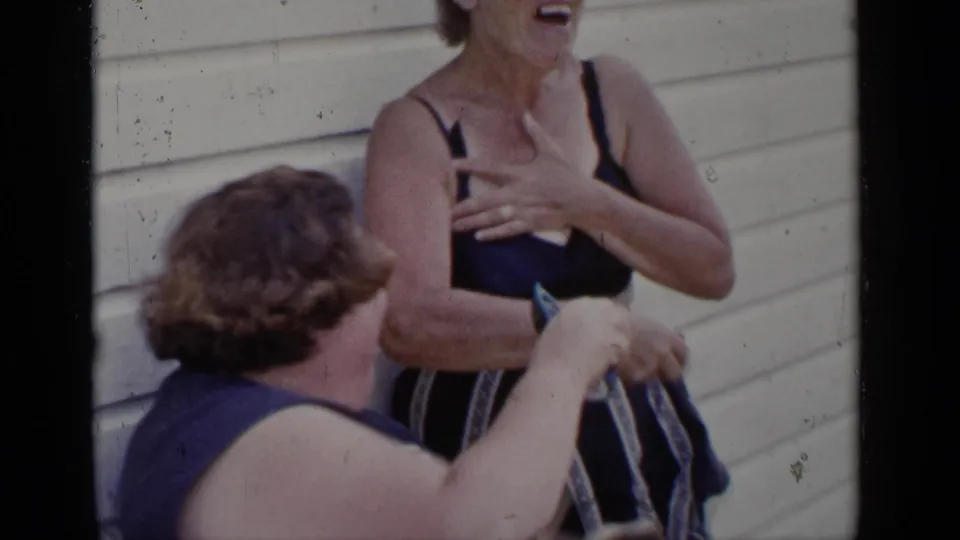 1951 New York 8mm frame of two older women laughing and gesturing outside, with swimwear and light house siding visible.