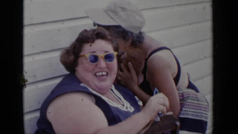 1951 New York 8mm frame with an older woman in yellow sunglasses smiling as a woman in a pale sun hat leans close beside her.