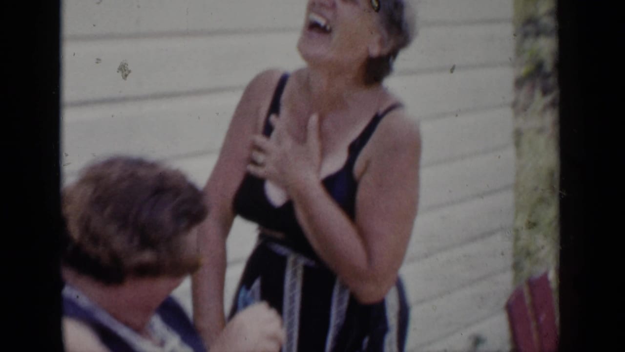 In 1951 New York home-movie footage, two women laugh beside a white-sided house, with one woman in a dark sleeveless garment leaning back.