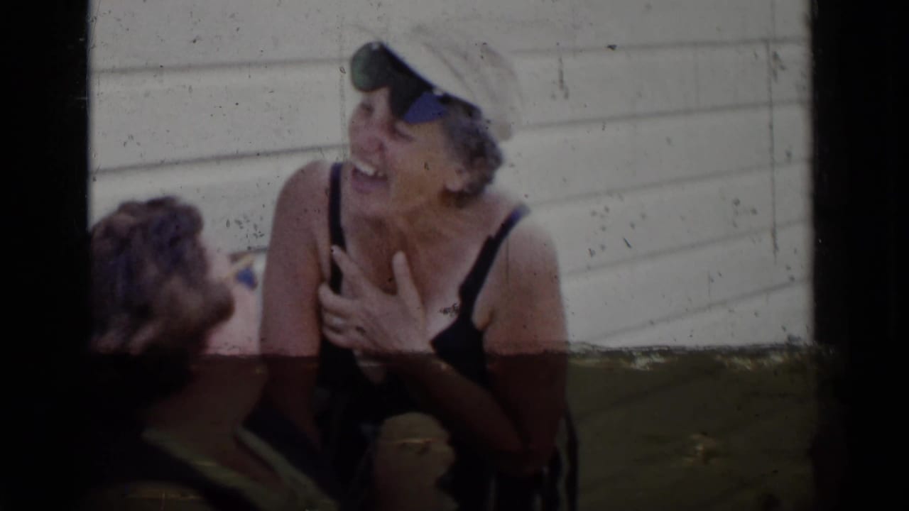 A woman in sunglasses and a light cap laughs beside another person near a white wall.