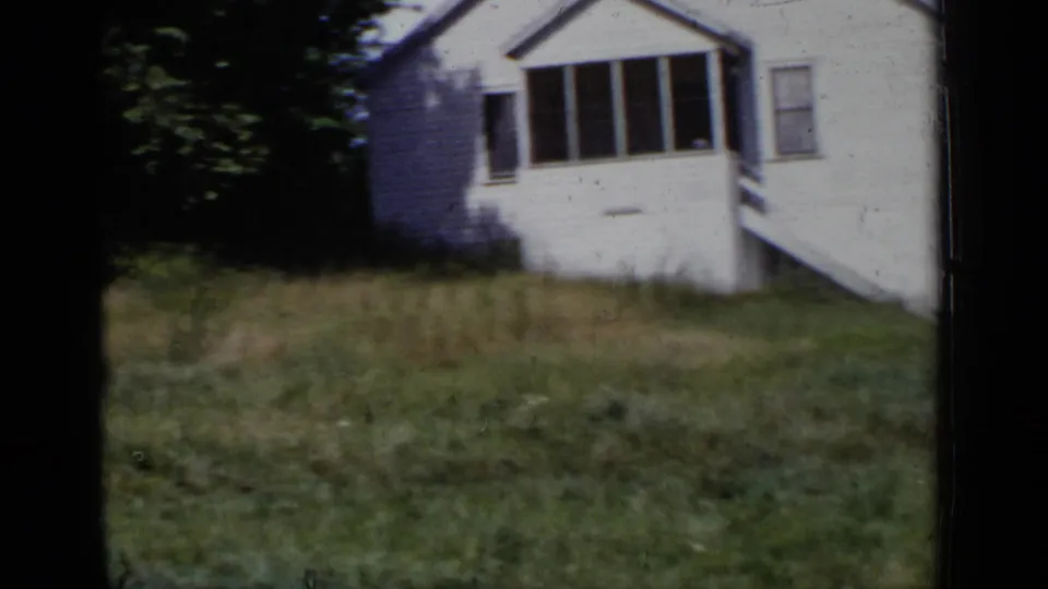 The camera turns to a white-sided house and grassy yard, showing porch windows and vegetation in the same New York home-movie setting.
