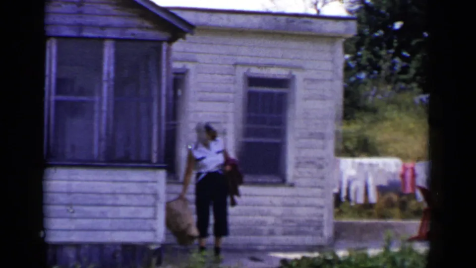 1951 New York home-movie frame showing an adult woman in a white shirt near a weathered white house and clothesline.