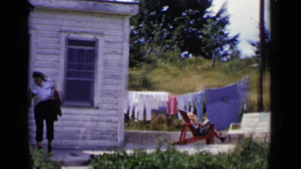1951 New York home-movie frame showing an adult woman beside a white house, with laundry, grass, and a red outdoor chair behind her.