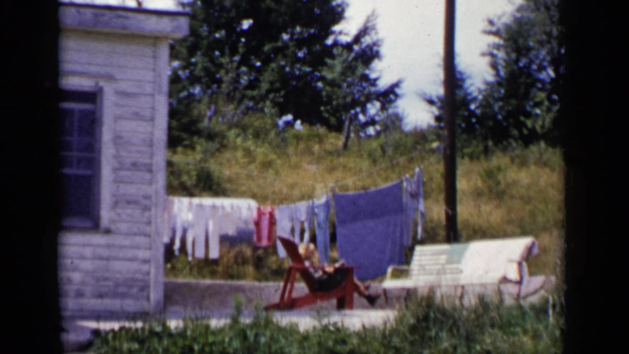 1951 New York home-movie frame showing a white clapboard house, laundry line, grassy yard, and a person resting near a red chair.