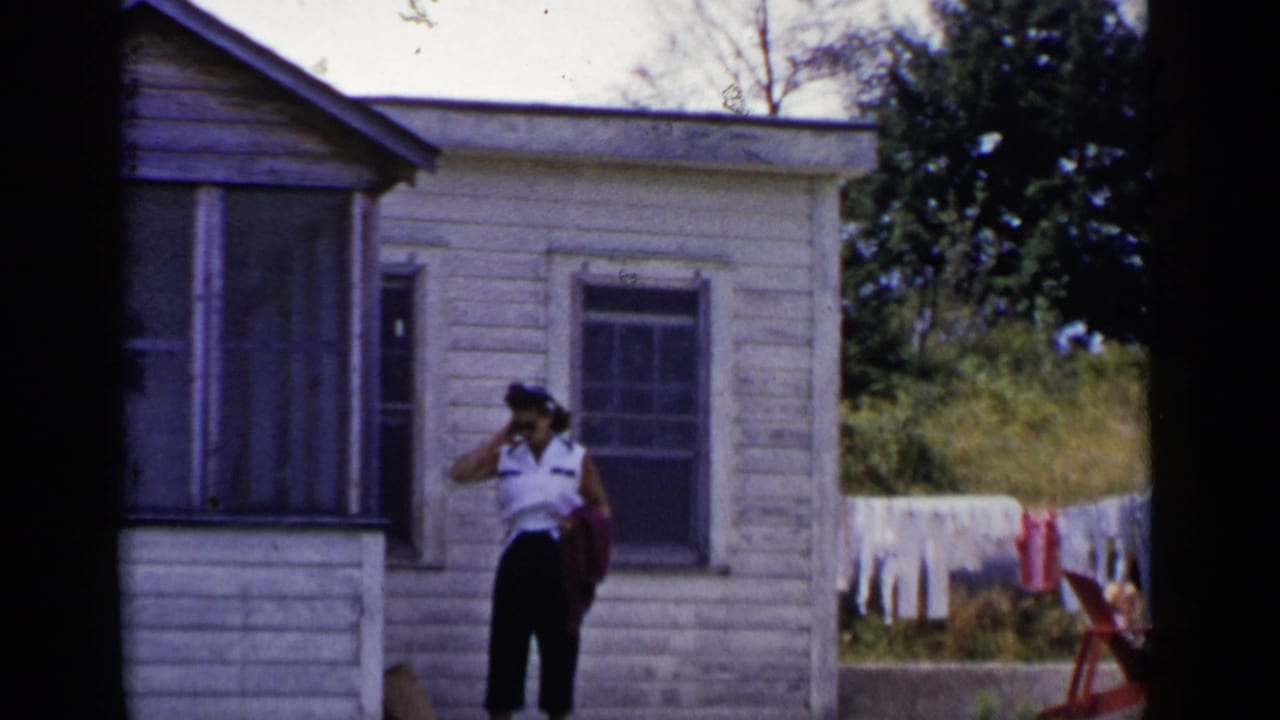 Woman in a white shirt stands near a weathered house with laundry behind her.