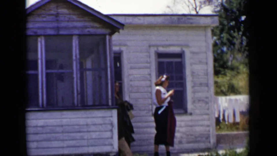 The woman appears to adjust her hat or hair beside the porch of the weathered white house in this 1951 New York home-movie frame.
