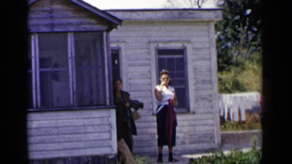 A woman faces the camera in front of the white house as another adult is partly visible in the doorway, with laundry in the yard.