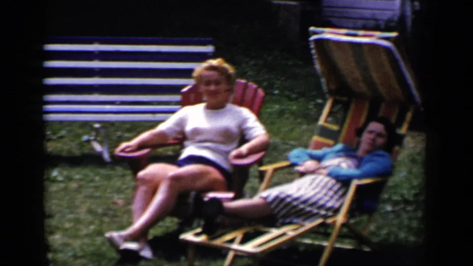 1951 New York home-movie frame showing two women relaxing in yard chairs, with a striped bench and grassy lawn visible behind them.
