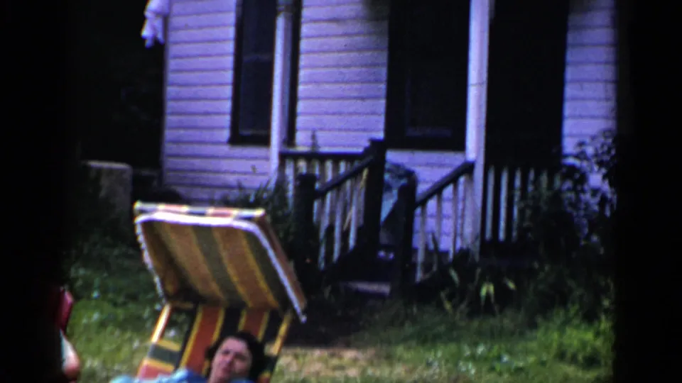 1951 New York frame showing a striped lounge chair in a yard near a house exterior, porch steps, railing, and siding.