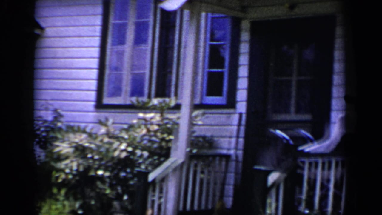 House facade with clapboard siding, tall windows, shrubs, and part of a porch railing.