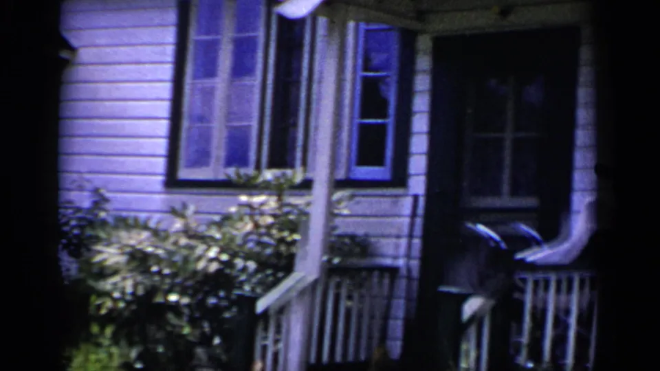 1951 New York exterior frame with white clapboard siding, dark-trimmed windows, dense shrubs, and a partial porch rail.