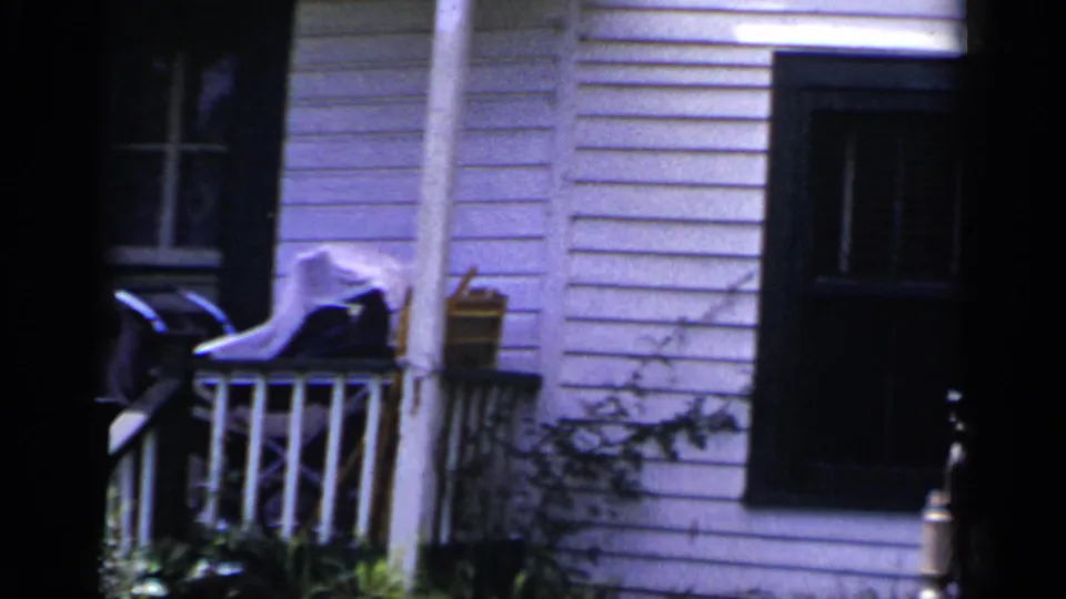 1951 New York home-movie view of a clapboard house corner with porch railing, dark window, downspout, and yard plants.