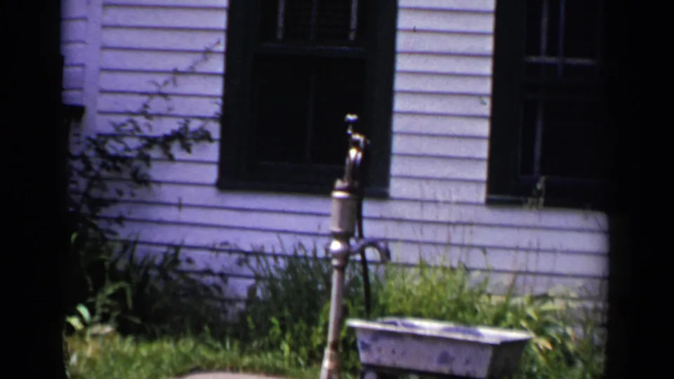 1951 New York exterior frame showing clapboard siding, dark windows, overgrown grass, shrubs, and a small metal yard fixture.