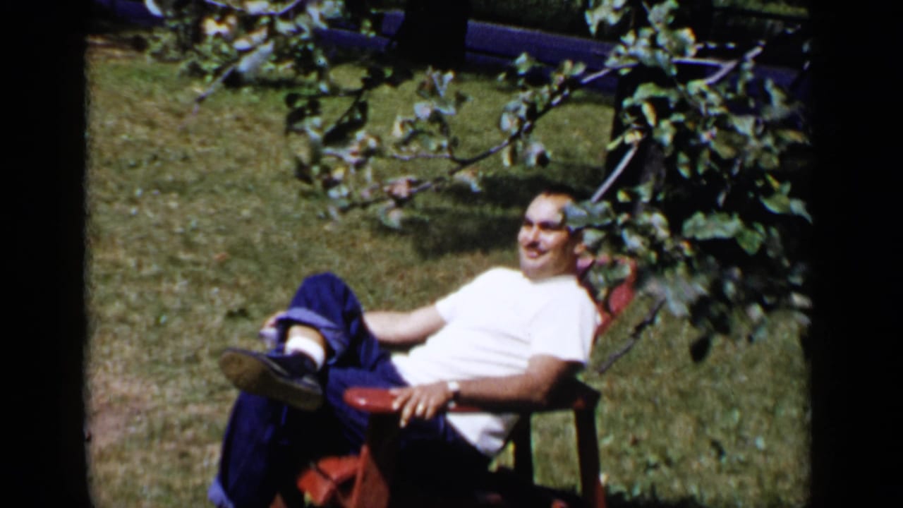 Man in a white shirt reclines in a red lawn chair under leafy trees.