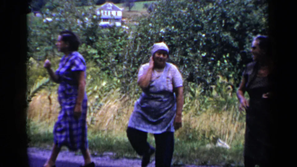 Two women in 1951 New York home-movie footage walk near tall grass and trees, with another adult partly entering at the right edge.