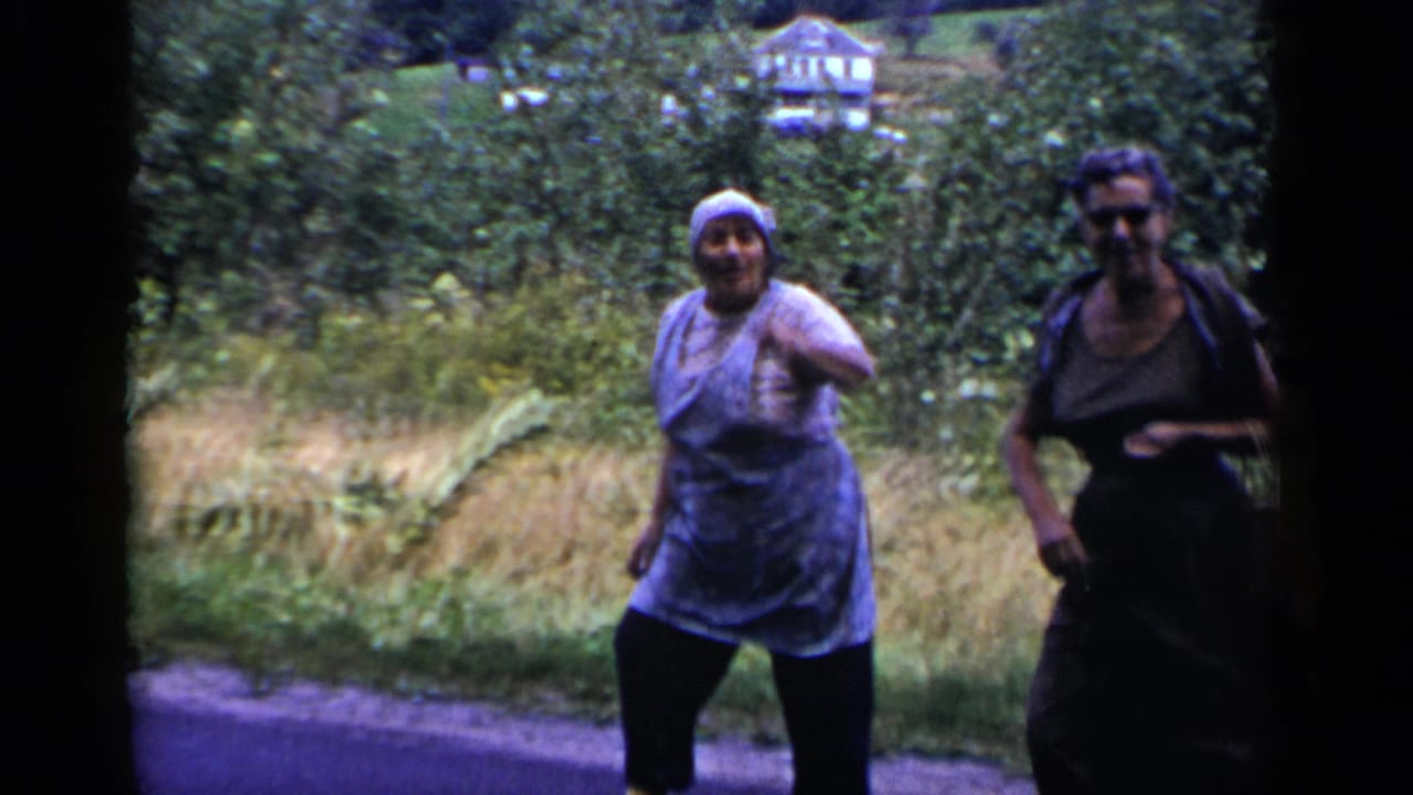 Two women walk along a roadside, one in a headscarf and apron and one in a dark dress.