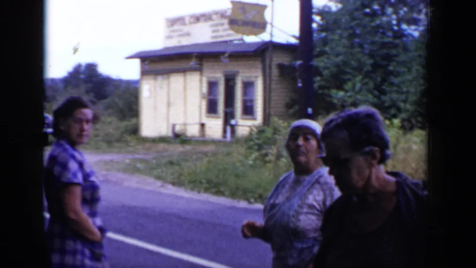 Three adult women are seen beside a rural New York road, with a small yellow roadside building and sign visible behind them.