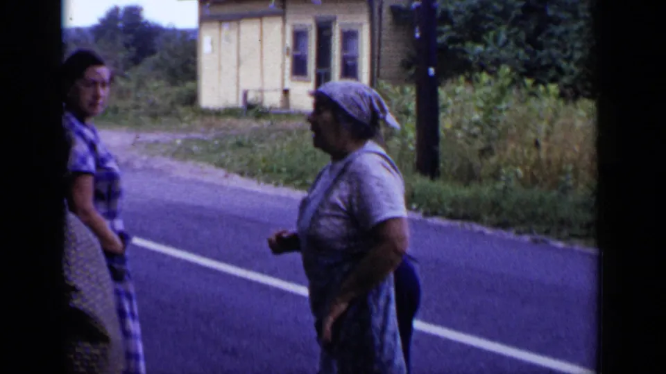 Two adult women face each other beside a rural New York road, with a small building, utility pole, and trees in the background.