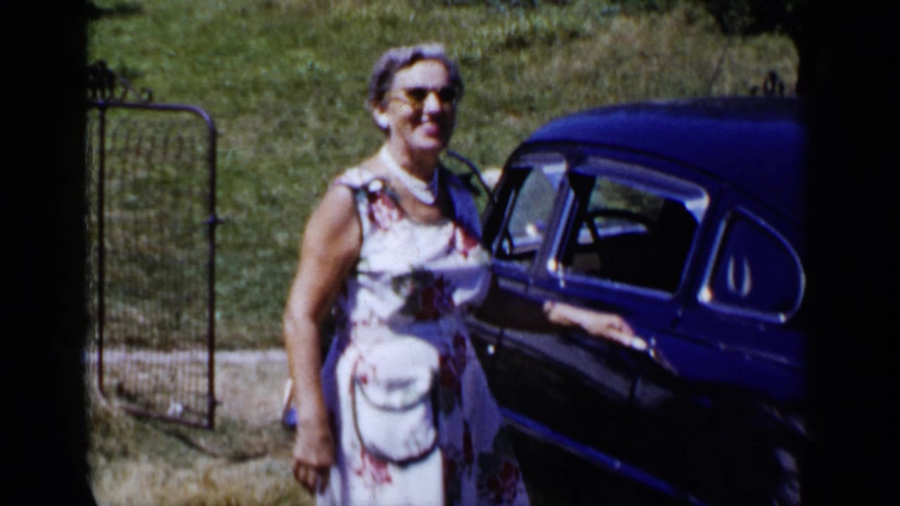 Woman in sunglasses and a floral dress standing beside a dark-blue vintage car.
