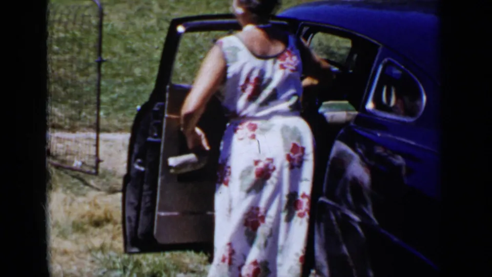Seen from behind, the woman in a floral dress steps toward the open door of the dark-blue car on a sunny grassy lot.