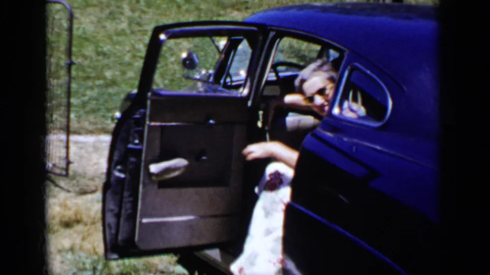 The woman sits inside the dark-blue car with the door open, looking out through sunglasses as sunlight falls on the grassy background.