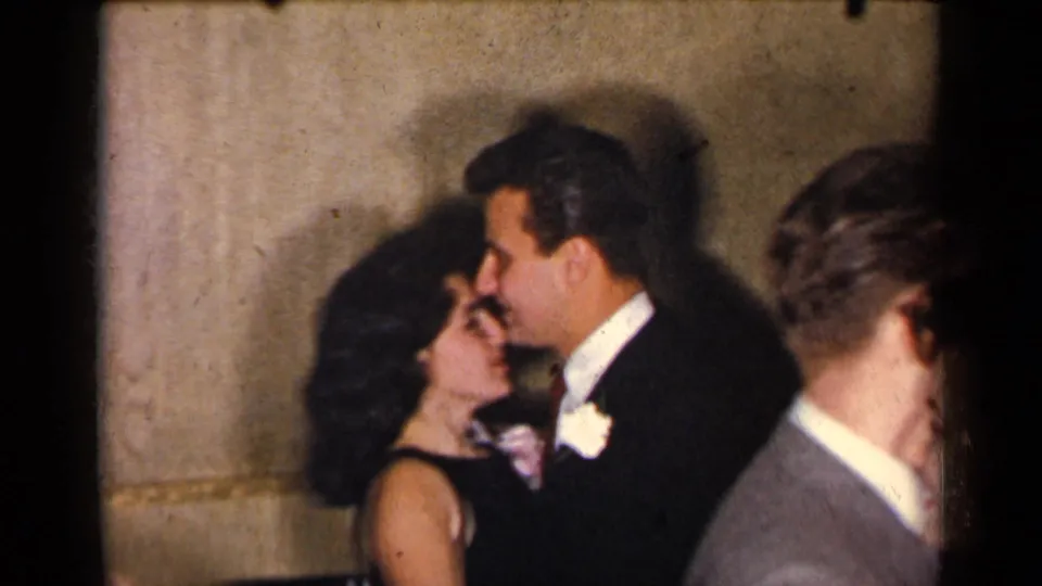 A formally dressed couple dances cheek-to-cheek in the supplied 1954 New York home-movie scene, with other guests partly visible.