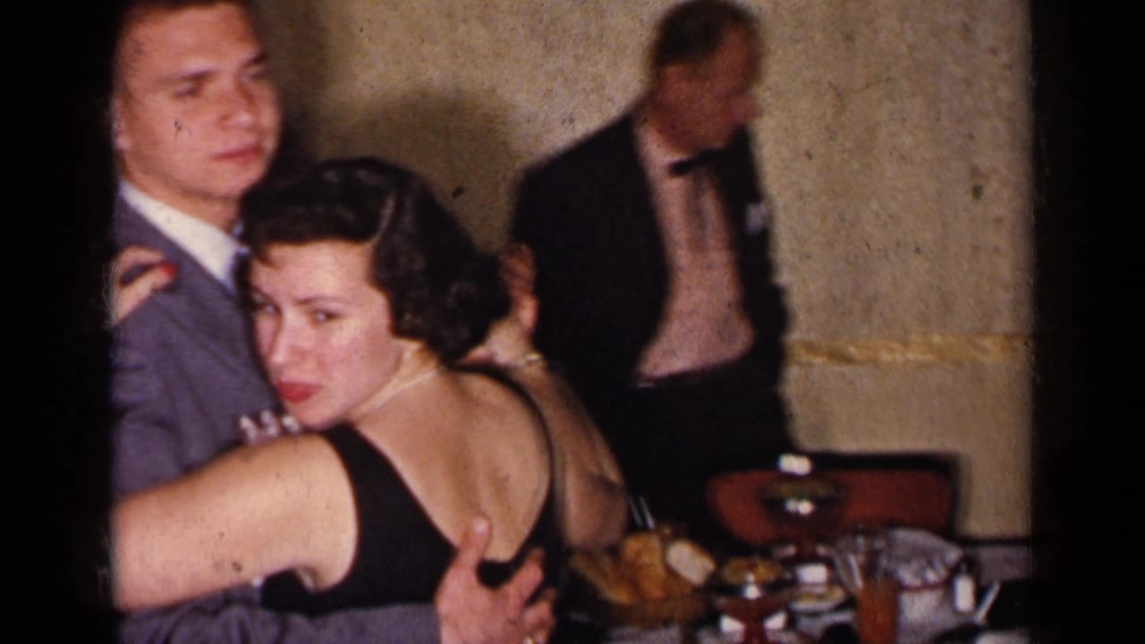 A woman in a low-back black dress looks toward the camera while dancing with a suited man at a 1950s New York indoor social.
