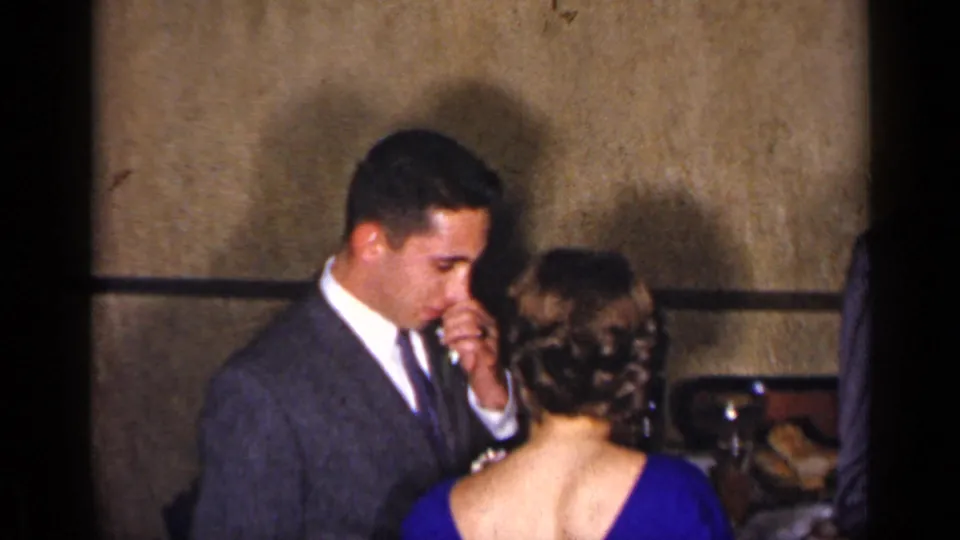 A suited man dances with a woman in a blue dress beside banquet table settings in supported 1954 New York home-movie footage.