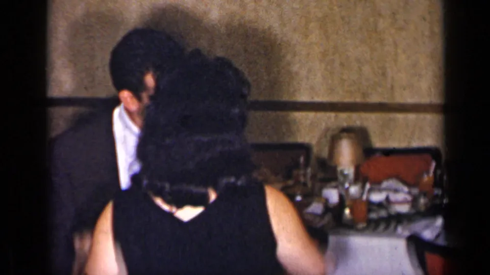 In a 1954 New York home-movie frame, a suited man and woman in a dark sleeveless dress dance near tables set for a gathering.