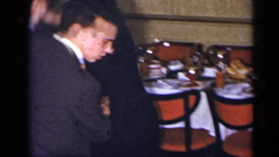 A man in glasses and a dark suit dances with a woman in a dim reception-like room, with plates, glasses, and red chairs visible.