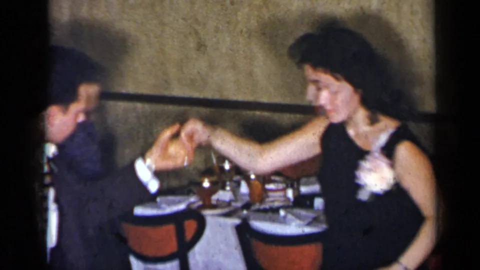A woman in a sleeveless dark dress holds her partner's hand while dancing in a 1954 New York interior with table settings behind them.