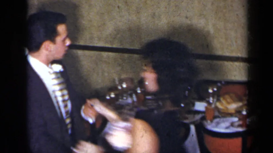 1954 New York home-movie frame of a suited man with boutonniere beside a woman in a black dress at an indoor dinner table.