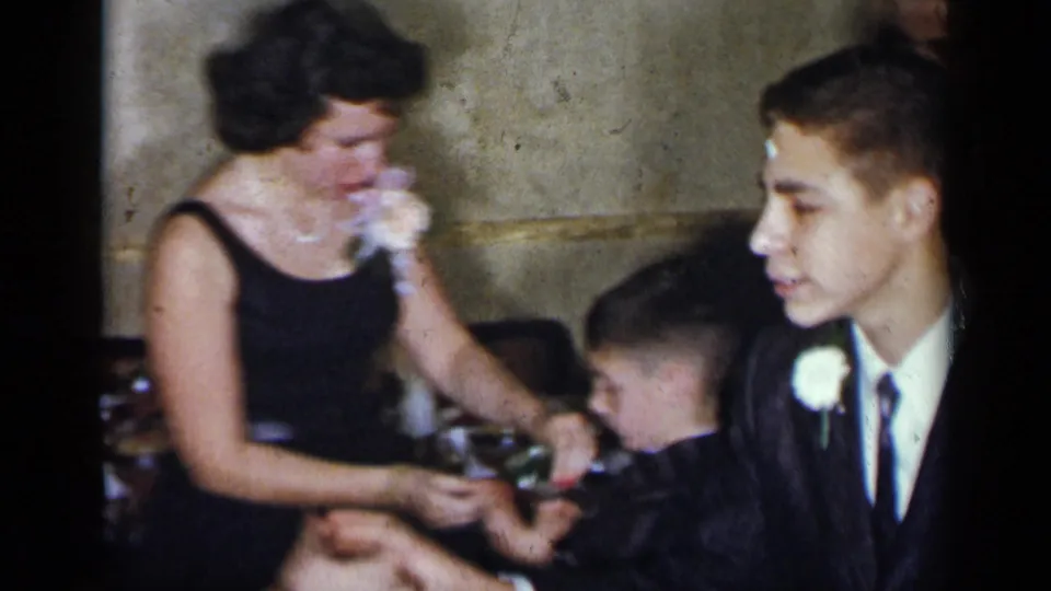 1954 New York home-movie frame showing a woman in a black dress seated at a table with a suited young male guest and another shorter male guest.