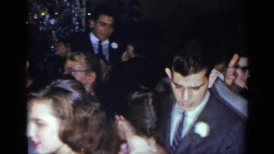 In 1954 New York home-movie footage, a young man in a dark suit and white boutonniere passes through guests at a crowded formal party.
