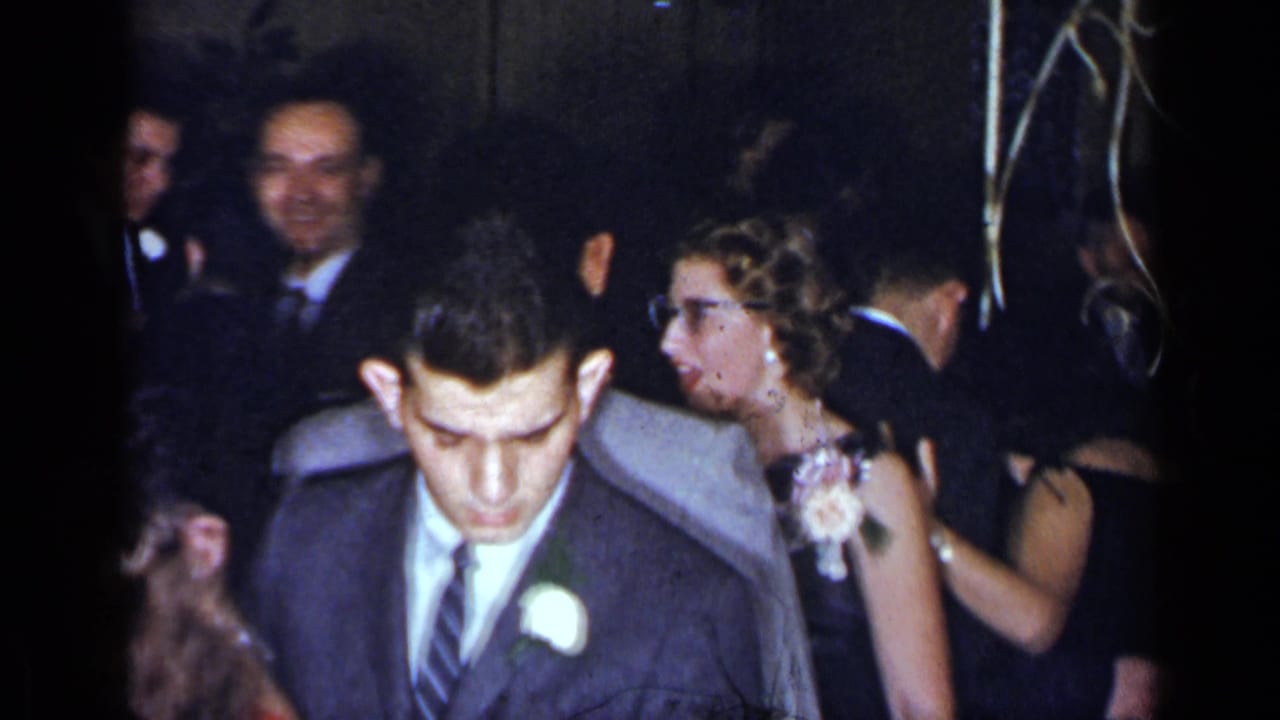 Young man with boutonniere looks downward while guests mingle behind him.