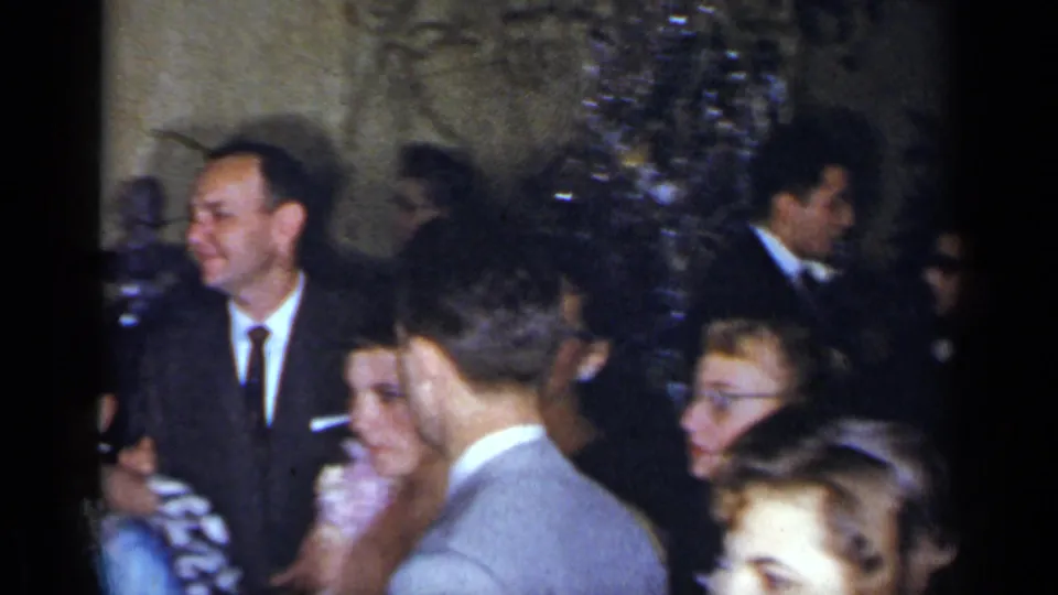 At the crowded 1954 New York gathering, formally dressed guests mingle in a dim interior while the handheld camera catches candid faces.