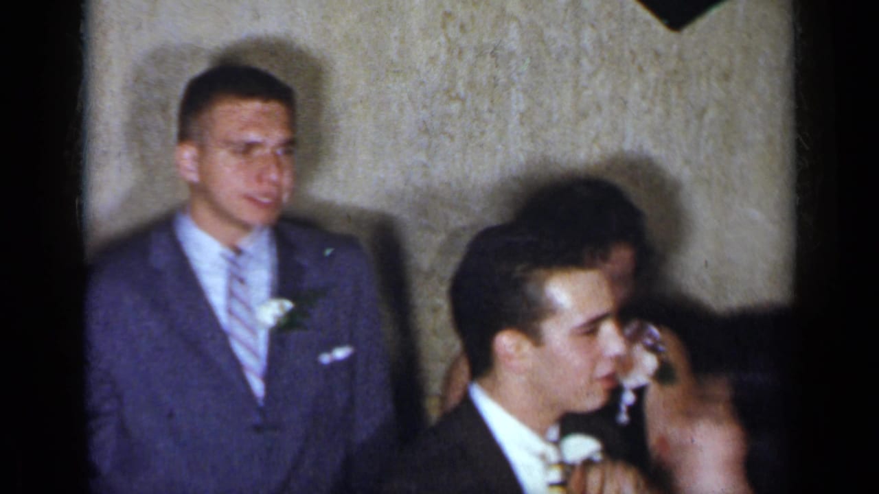 Suited young men stand near a woman at a dim formal indoor gathering.