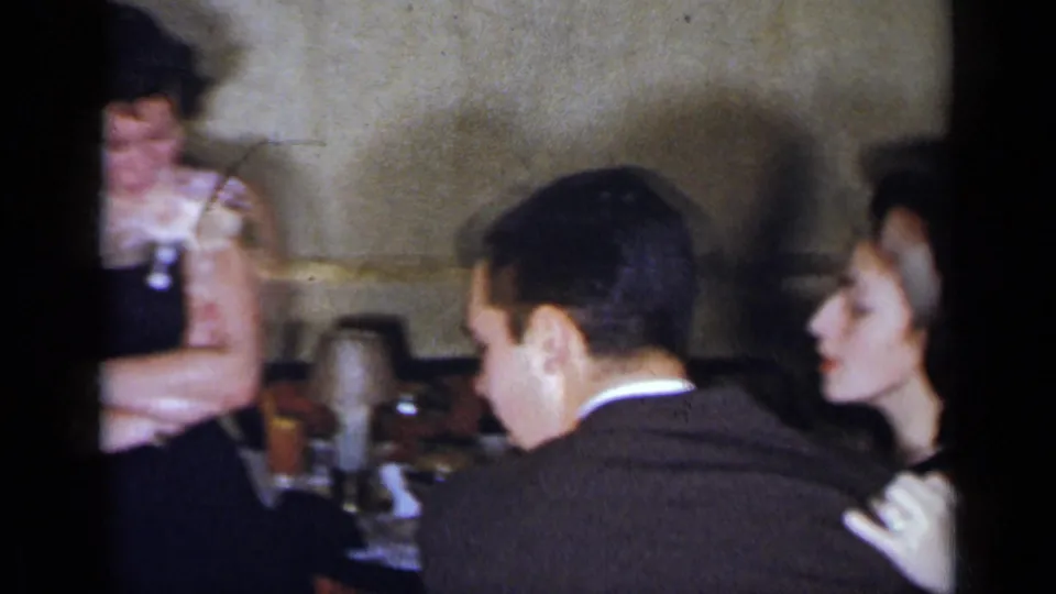 1954 New York home-movie frame with a young couple near tables and chairs during a formal indoor dance or social event.