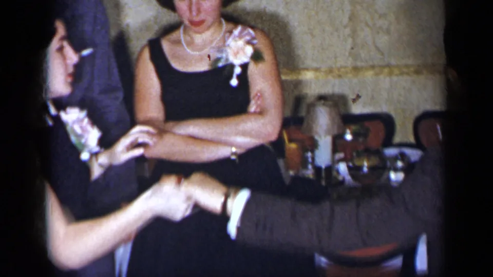 1954 New York frame showing women in dark formal dresses with corsages near a suited man at an indoor social gathering.