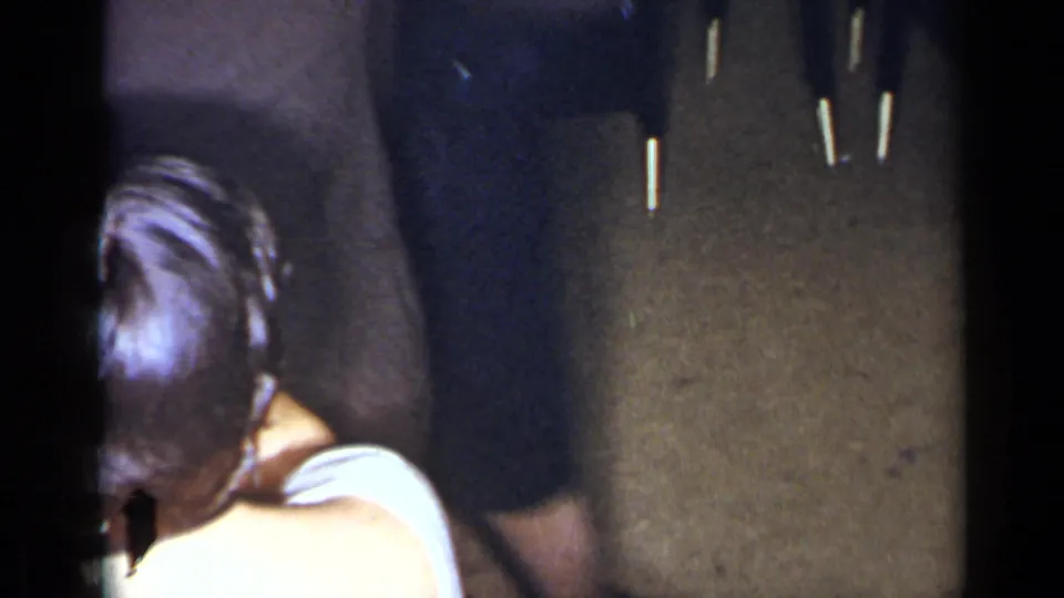 1954 New York party frame with adults in dark clothing and a person in a light top partly turned away indoors.