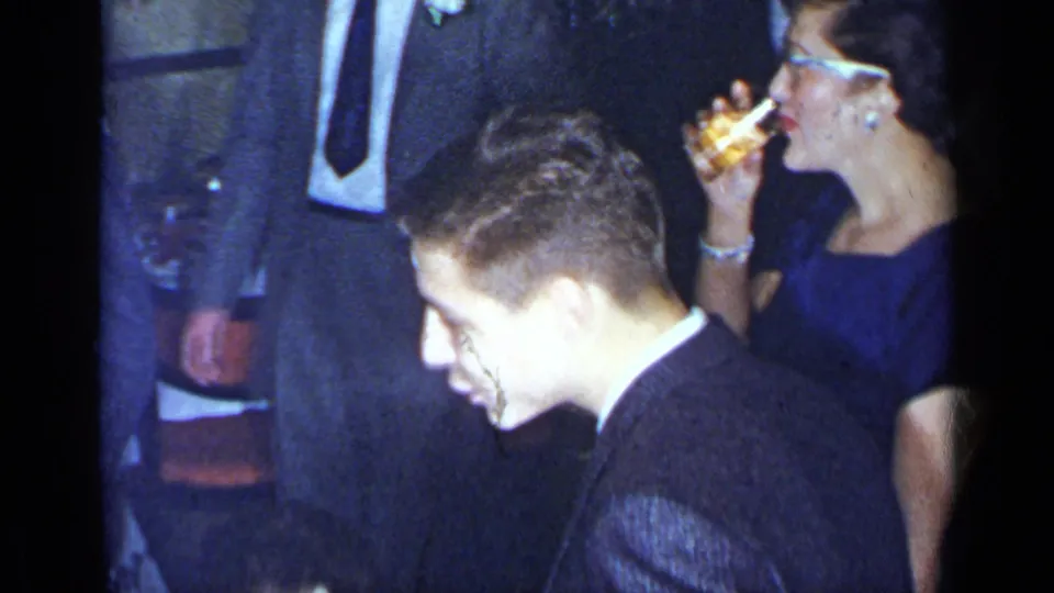 1954 New York party frame with a woman in a blue dress drinking from a glass near a man in a dark suit.