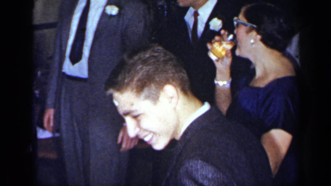Formal party guests mingle while a young man smiles in the foreground.