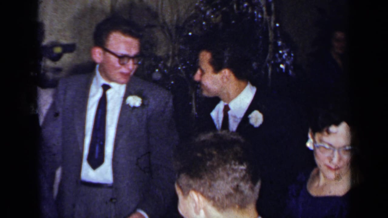 1954 New York party frame showing suited men with boutonnieres in conversation among guests in a dim indoor room.