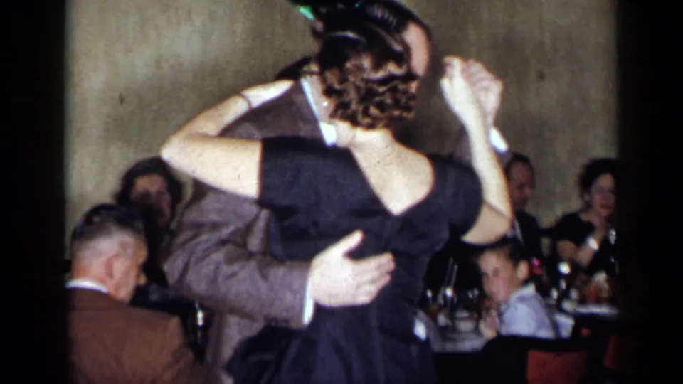 1954 New York formal party frame with a couple dancing in the foreground and seated guests at tables behind them.
