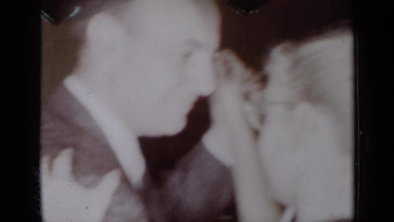 Two adults in soft focus lean close together in a dim indoor home-movie frame.