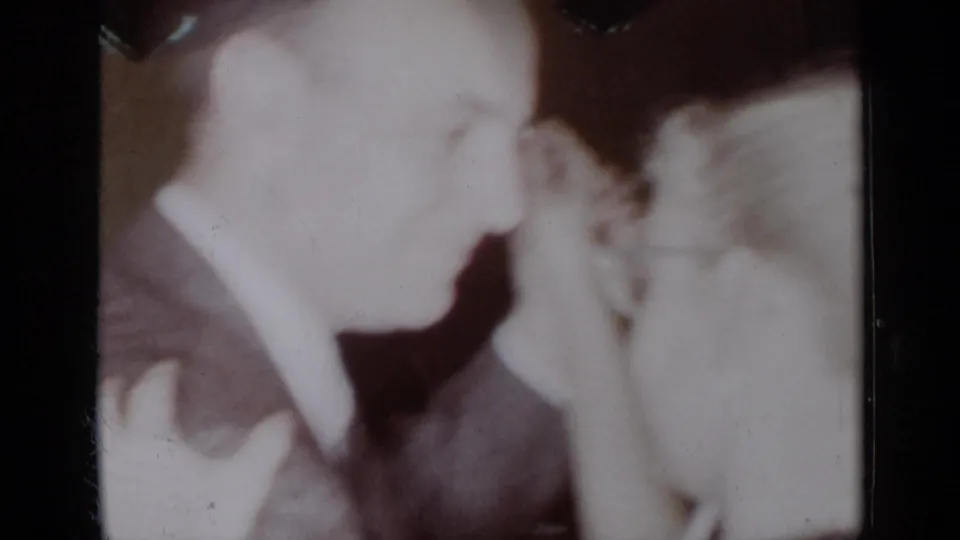 In this 1954 New York 8mm frame, a man in a suit and a light-haired woman appear close together in a dim private interior.